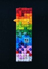 A vertically arranged rectangular structure made out of colorful LEGO bricks, featuring a wide range of colors including red, orange, yellow, green, blue, and pink. There is a small graphic in the middle of the composition displaying text and stylized faces. The background is a solid dark color, which contrasts with the vivid hues of the bricks.