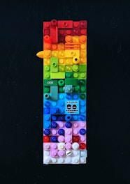A vertically arranged rectangular structure made out of colorful LEGO bricks, featuring a wide range of colors including red, orange, yellow, green, blue, and pink. There is a small graphic in the middle of the composition displaying text and stylized faces. The background is a solid dark color, which contrasts with the vivid hues of the bricks.