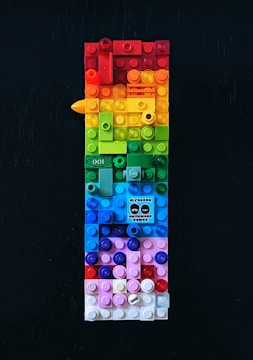 A vertically arranged rectangular structure made out of colorful LEGO bricks, featuring a wide range of colors including red, orange, yellow, green, blue, and pink. There is a small graphic in the middle of the composition displaying text and stylized faces. The background is a solid dark color, which contrasts with the vivid hues of the bricks.