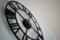 A decorative wall clock with a minimalist design, featuring Roman numerals and slender gold hands. The clock has a black, open-frame structure, showcasing a modern aesthetic against a light wall.