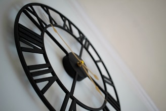 A decorative wall clock with a minimalist design, featuring Roman numerals and slender gold hands. The clock has a black, open-frame structure, showcasing a modern aesthetic against a light wall.