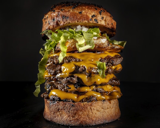 A towering cheeseburger stacked with multiple beef patties, each layered with melting cheddar cheese. It is topped with crispy lettuce, sliced onions, and possibly fried onions. The bun is seeded and appears slightly toasted.