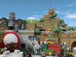 A vibrant and playful theme park scene with structures resembling blocks, coins, and characters from a popular video game franchise. The area features a castle with menacing stone statues, tiered earthwork adorned with cartoonish elements like question blocks and smiling clouds. Various colorful pipes, oversized mushrooms, and playful characters such as piranha plants are scattered throughout. Visitors are walking around, enjoying the themed environment.