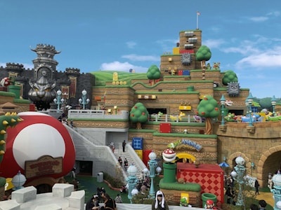 A vibrant and playful theme park scene with structures resembling blocks, coins, and characters from a popular video game franchise. The area features a castle with menacing stone statues, tiered earthwork adorned with cartoonish elements like question blocks and smiling clouds. Various colorful pipes, oversized mushrooms, and playful characters such as piranha plants are scattered throughout. Visitors are walking around, enjoying the themed environment.
