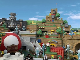 A vibrant and playful theme park scene with structures resembling blocks, coins, and characters from a popular video game franchise. The area features a castle with menacing stone statues, tiered earthwork adorned with cartoonish elements like question blocks and smiling clouds. Various colorful pipes, oversized mushrooms, and playful characters such as piranha plants are scattered throughout. Visitors are walking around, enjoying the themed environment.