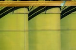 Three large green storage tanks feature prominently, with yellow pipes running horizontally across the top portion. Shadows cast by the pipes create diagonal patterns on the tanks, adding depth and contrast.