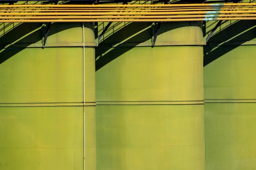 Three large green storage tanks feature prominently, with yellow pipes running horizontally across the top portion. Shadows cast by the pipes create diagonal patterns on the tanks, adding depth and contrast.