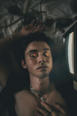 A person with closed eyes is lying down with orange objects covering their eyelids. They are wearing multiple silver chains and rings, and their hand rests on their chest. The individual is near a screen emitting light. The background appears to be a bed with some indistinct items.