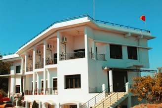 A modern, two-story building painted in white with multiple windows and a staircase leading to the entrance. The structure has black railings and air conditioning units installed on the upper level. A red flag is flying from the rooftop, and there are trees and shrubs around the building. The sky is clear and blue.