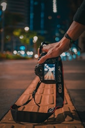 A hand holding a DSLR camera with a visible camera strap reading 'EOS DIGITAL'. The camera is positioned above a sidewalk with textured tiles, capturing a street scene with bokeh lights in the background. The atmosphere suggests it's nighttime, with blurred streetlights and some greenery visible from a distance.