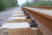 A close-up view of a railway track extends into the distance. The focus is on the metal rail and fastening system on the concrete sleepers, highlighting the texture and materials.