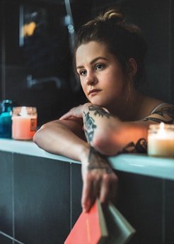 A person with tattoos is relaxing in a dimly lit bath, surrounded by lit candles. They have a thoughtful expression and are holding a book in one hand, which rests on the edge of the bathtub. The room has a serene and introspective atmosphere.