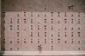 A wall composed of multiple small, marble plaques, each engraved with an emblem and inscription. Some plaques display small, decorative symbols and arrangements of letters, while others are marked with crosses and floral elements. A single bouquet of flowers rests at the bottom right corner.