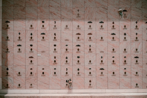A wall composed of multiple small, marble plaques, each engraved with an emblem and inscription. Some plaques display small, decorative symbols and arrangements of letters, while others are marked with crosses and floral elements. A single bouquet of flowers rests at the bottom right corner.