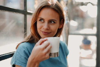 A smiling woman enjoying a peaceful moment with a cup of herbal tea by a sunny window.