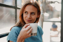 A woman smiling gently while holding a cup of herbal tea near a sunny window.