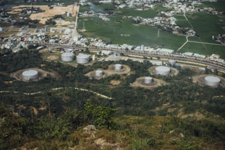 An aerial view showcases several large industrial storage tanks surrounded by lush greenery. In the background, a residential area with numerous small buildings and houses is visible, along with expanses of cultivated fields.