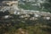 An aerial view showcases several large industrial storage tanks surrounded by lush greenery. In the background, a residential area with numerous small buildings and houses is visible, along with expanses of cultivated fields.
