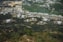 An aerial view showcases several large industrial storage tanks surrounded by lush greenery. In the background, a residential area with numerous small buildings and houses is visible, along with expanses of cultivated fields.