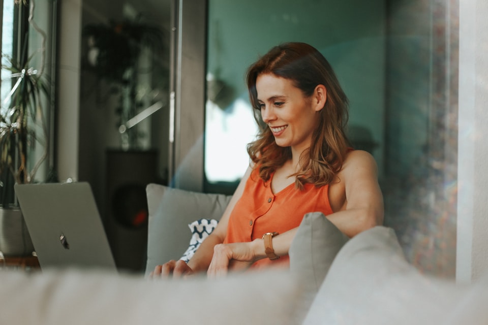 Woman sitting with beige cushions and smiling while using a laptop.