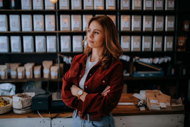 A woman with long hair and a watch on her wrist stands confidently with arms crossed in a shop with shelves filled with various labeled bags and containers. The setting is warm and inviting, with subtle lighting adding to the cozy atmosphere.