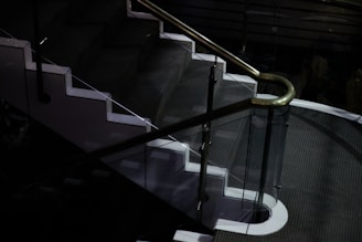 A dimly lit staircase with a sleek design, featuring glass panels and white edges, complemented by a metallic handrail. The surrounding area is dark, highlighting the stairway.