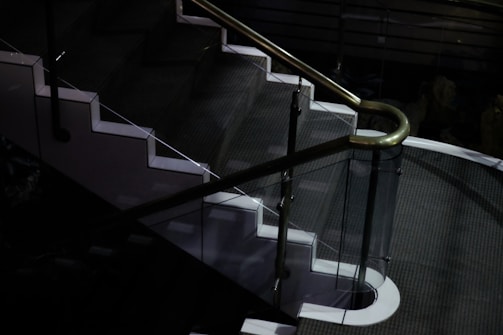 A dimly lit staircase with a sleek design, featuring glass panels and white edges, complemented by a metallic handrail. The surrounding area is dark, highlighting the stairway.