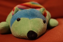 A colorful stuffed toy that resembles an animal, possibly a hedgehog, lying on a brown surface. The toy is predominantly green with a blue face, black eyes, and nose. It has a multi-colored, rainbow-patterned back.