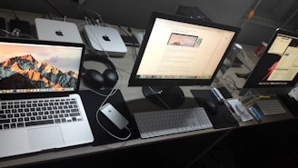 A neatly arranged workspace featuring three computer screens, including a MacBook, an iMac, and a second monitor. On the desk, there's a pair of headphones, a mouse, keyboards, a notepad, and other office items. In the background, two Mac Mini units and other electronic accessories are visible.