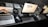 A neatly arranged workspace featuring three computer screens, including a MacBook, an iMac, and a second monitor. On the desk, there's a pair of headphones, a mouse, keyboards, a notepad, and other office items. In the background, two Mac Mini units and other electronic accessories are visible.