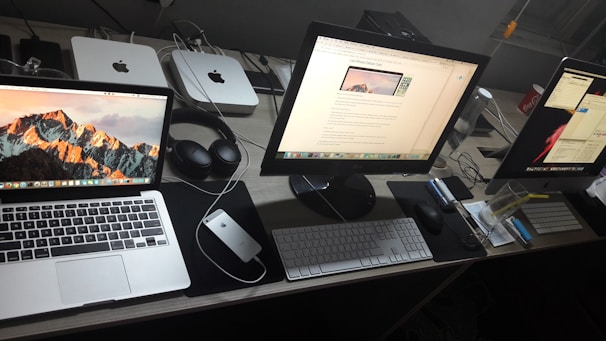 A neatly arranged workspace featuring three computer screens, including a MacBook, an iMac, and a second monitor. On the desk, there's a pair of headphones, a mouse, keyboards, a notepad, and other office items. In the background, two Mac Mini units and other electronic accessories are visible.