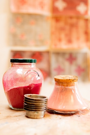 Two glass jars, one filled with vibrant red liquid and another with pink powder, are placed on a wooden surface. A stack of small metal cups stands nearby. The background features out-of-focus patterned squares in warm, earthy colors.