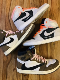 Four high-top sneakers arranged in an alternating pattern on a wooden surface. Two shoes have pink laces and two have orange laces. The shoes predominantly feature black swooshes, white panels, and shades of gray or brown sections.