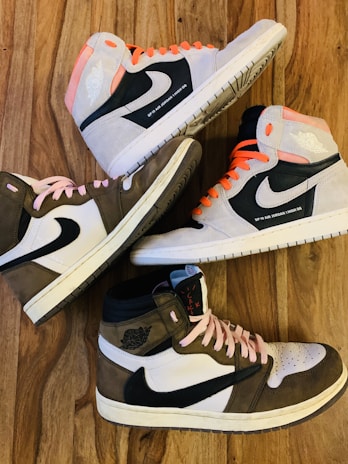 Four high-top sneakers arranged in an alternating pattern on a wooden surface. Two shoes have pink laces and two have orange laces. The shoes predominantly feature black swooshes, white panels, and shades of gray or brown sections.