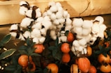Cotton bolls with fluffy white fibers are growing alongside small orange fruits and dark green leaves, set against a wooden background.