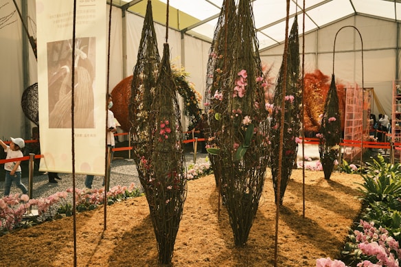 Inside a large, well-lit exhibition tent, several vertical, elongated structures made of woven natural materials are decorated with pink flowers. The ground is covered in mulch, and there are various potted plants and flowers around. People are visible in the background, with some observing the displays. A large banner with text and an image hangs on the left side, while orange barricades guide visitors through the space.