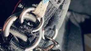 A close-up view of heavy-duty metal rings and thick, durable fabric straps, likely part of a harness or industrial equipment. The lighting creates a stark contrast, highlighting the textures and metallic surfaces.