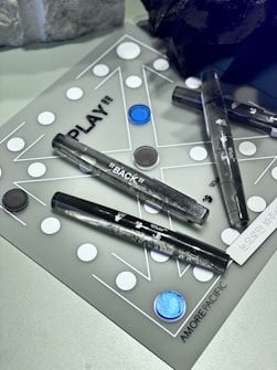 A board game layout featuring a combination of black and white circular pieces distributed across a translucent surface with geometric lines. The space includes several clear acrylic tubes with inscriptions, and a labeled section that reads 'AMORE PACIFIC'. A few irregularly shaped dark and light objects are placed alongside the game setup.