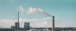A large industrial facility with two prominent smokestacks emitting white smoke into a blue sky. Numerous power lines and electrical towers stretch across the landscape in the background. The area is expansive, with buildings and infrastructure indicating a power plant or manufacturing site.
