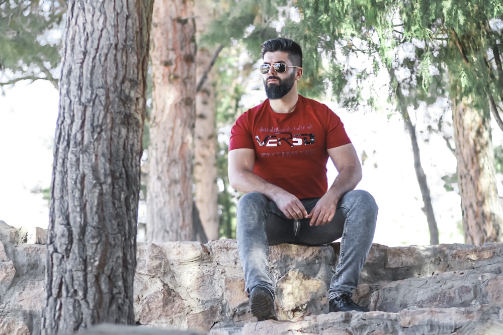 man in red crew neck t-shirt and blue denim jeans sitting on brown tree log