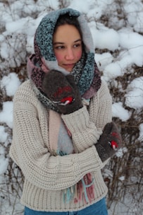 A person stands outdoors in a snowy environment, wearing a thick knitted sweater and a multi-colored scarf wrapped around their head and neck. They have on brown mittens featuring a small red and white pattern. The background consists of snow-covered branches.