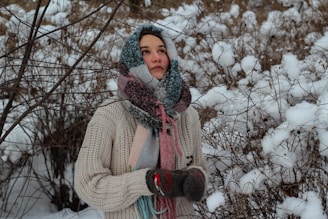 A person wearing a thick knitted sweater, colorful scarf, and gloves stands amidst snowy branches and a wintery landscape. The person looks thoughtful, with their hands clasped together as if trying to stay warm.
