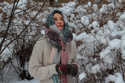 A person wearing a thick knitted sweater, colorful scarf, and gloves stands amidst snowy branches and a wintery landscape. The person looks thoughtful, with their hands clasped together as if trying to stay warm.