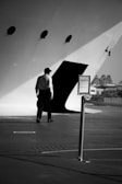 A person in formal attire is walking towards an entrance on a large vessel or ship. The setting is monochromatic and the lighting creates dramatic shadows. There is a signpost in the foreground and the background features some distant buildings.