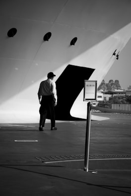 A person in formal attire is walking towards an entrance on a large vessel or ship. The setting is monochromatic and the lighting creates dramatic shadows. There is a signpost in the foreground and the background features some distant buildings.