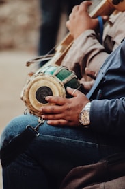 A person is holding a small, round percussion instrument with a wooden body and green finish, likely a type of drum. The individual's hand rests on the drum, and they are wearing a wristwatch. The person is dressed in casual attire, including a blue garment. The setting appears to be informal and relaxed.