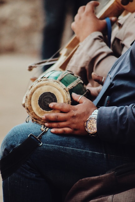 A person is holding a small, round percussion instrument with a wooden body and green finish, likely a type of drum. The individual's hand rests on the drum, and they are wearing a wristwatch. The person is dressed in casual attire, including a blue garment. The setting appears to be informal and relaxed.
