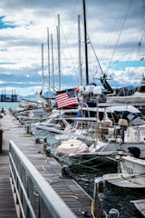 A marina with several sailboats docked along a wooden pier. The boats are equipped with masts and some have outdoor seating areas. An American flag is prominently displayed on one of the boats. The sky is partially cloudy with hints of blue peeking through. In the background, industrial buildings and mountains are visible.