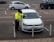 A parking enforcement officer wearing a high-visibility jacket is placing a ticket on a parked white Volkswagen car in a parking lot. The license plate of the car is visible, and several other cars are parked in the background.