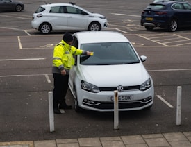 A parking enforcement officer wearing a high-visibility jacket is placing a ticket on a parked white Volkswagen car in a parking lot. The license plate of the car is visible, and several other cars are parked in the background.